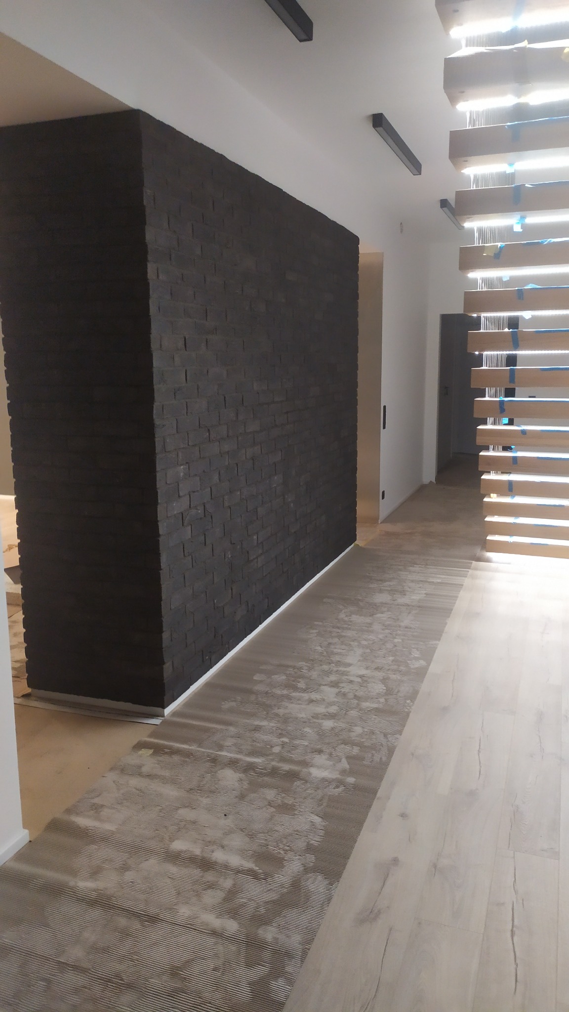 Ściana wykończona czarną cegłą dekoracyjną, widoczne ślady po pracach wykończeniowych na podłodze, w tle ażurowa konstrukcja schodów z zabezpieczeniami.