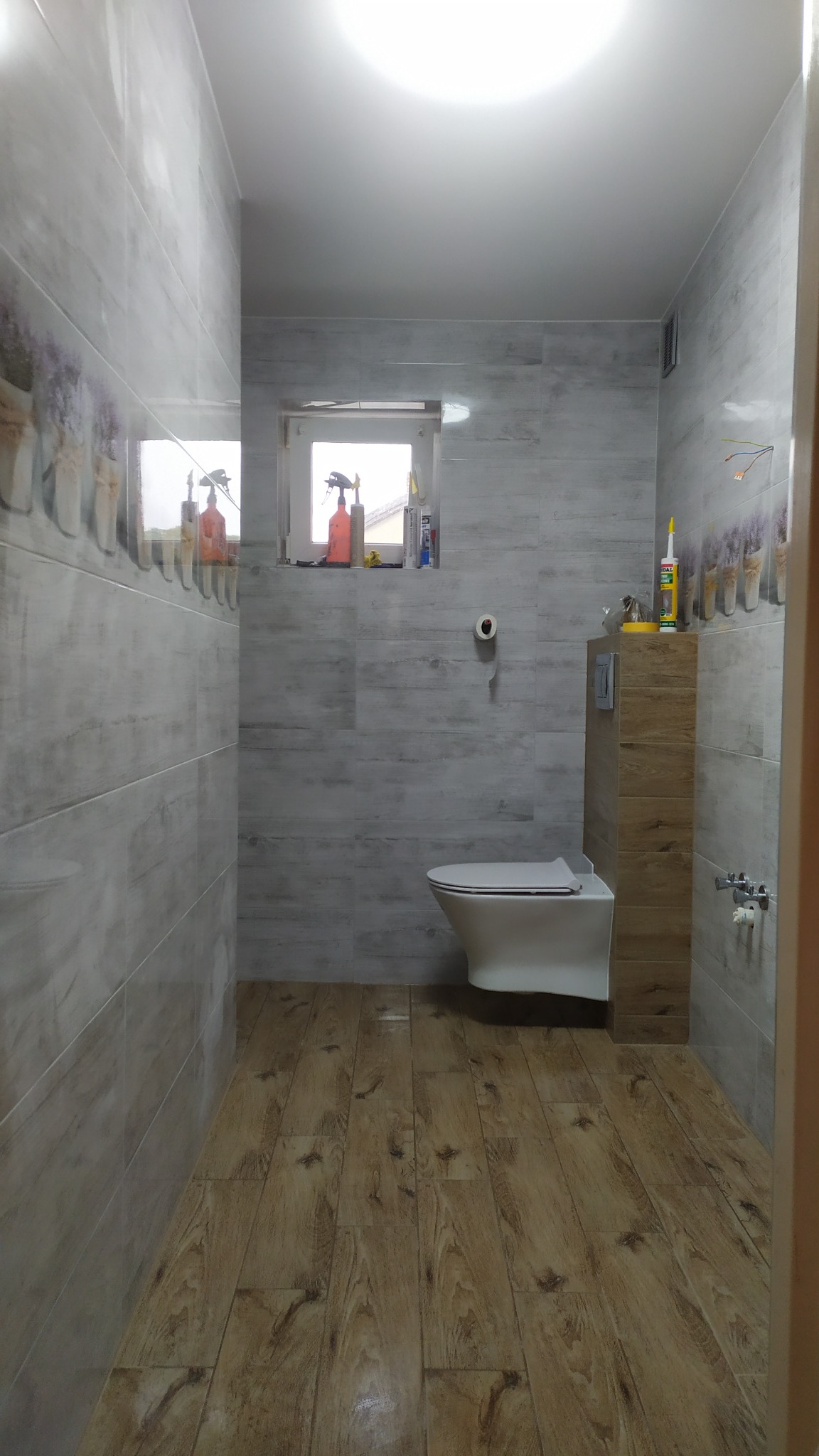 Wykończona łazienka z drewnopodobną podłogą, szarymi płytkami na ścianach z motywem lawendy, wiszącą toaletą i akcesoriami na parapecie okna.