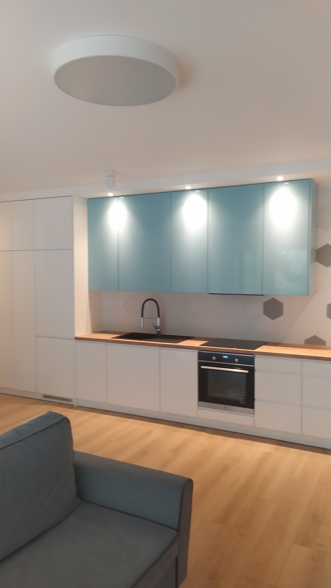 Nowoczesna kuchnia z białymi szafkami dolnymi i turkusowymi górnymi, drewnianym blatem, czarnym zlewem i płytą indukcyjną, oświetlona punktowo, z widocznym fragmentem szarej sofy.