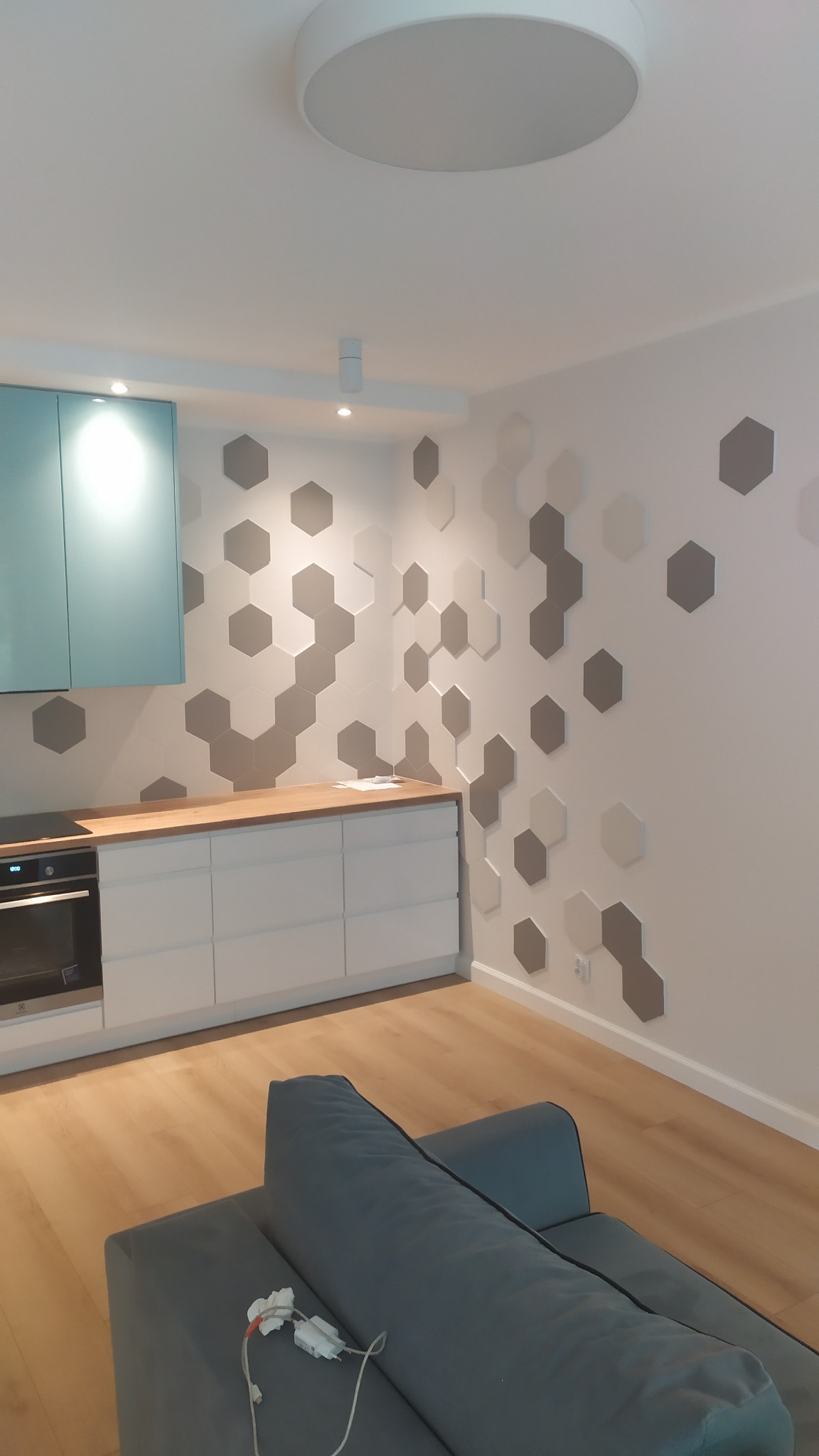 Nowoczesna kuchnia z białymi szafkami i drewnianym blatem, ściana ozdobiona szarymi i białymi heksagonami, widok z perspektywy salonu z szarą sofą i widocznym kablem zasilającym.