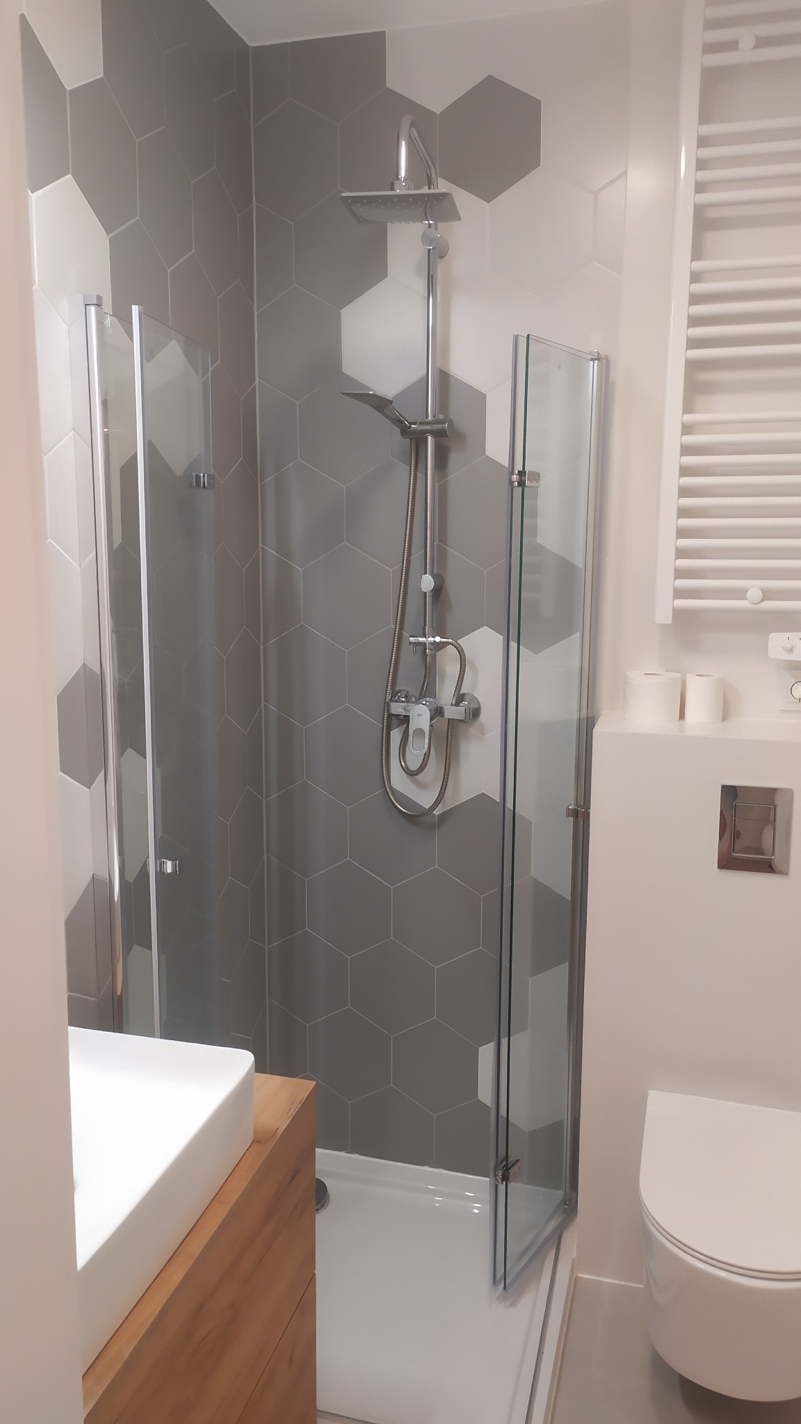 Nowoczesna łazienka z kabiną prysznicową typu walk-in, heksagonalnymi szaro-białymi płytkami na ścianach, deszczownicą i umywalką na drewnianej szafce.