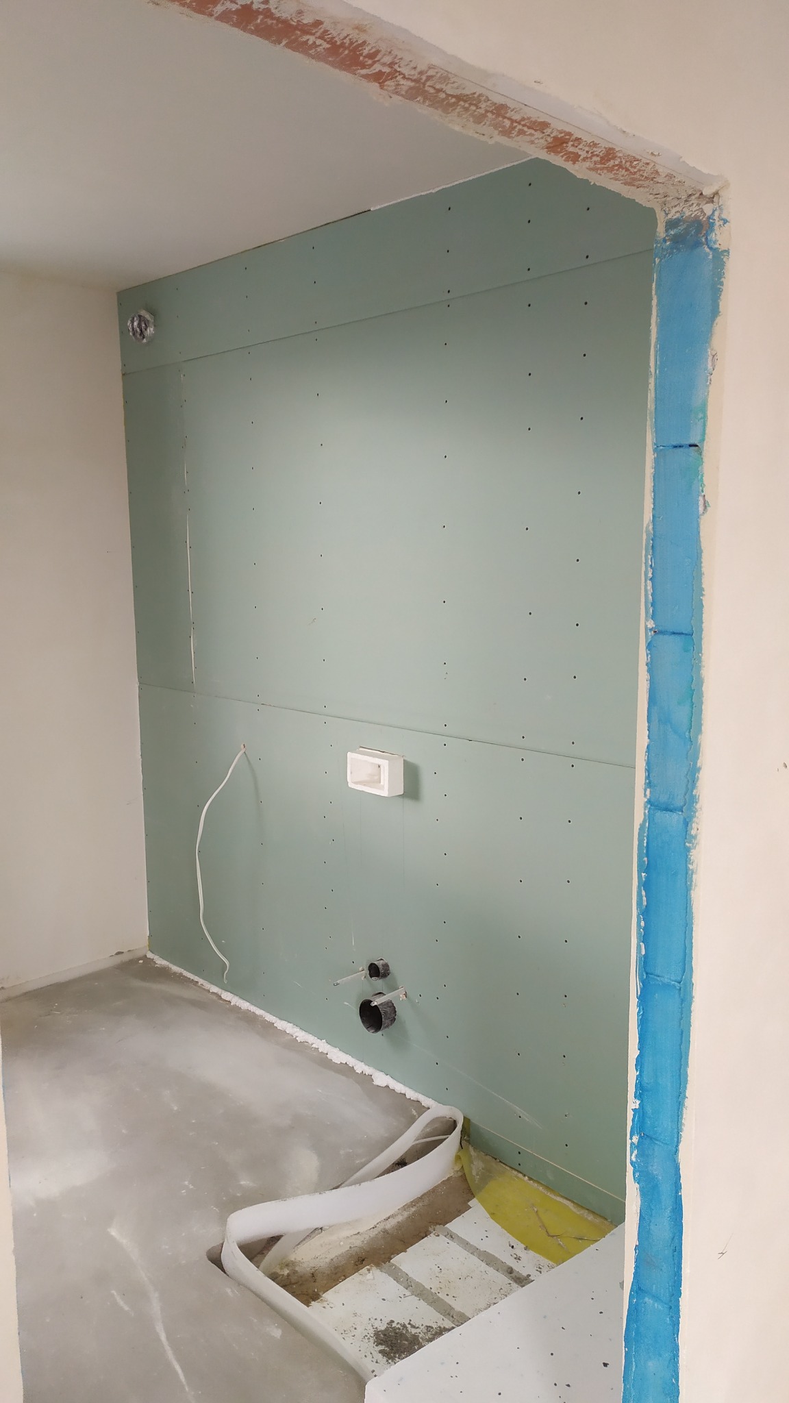 Ściana w trakcie montażu płyt gipsowo-kartonowych z widocznymi otworami na instalacje, surowy beton na podłodze i zabezpieczone niebieską taśmą framugi drzwi.