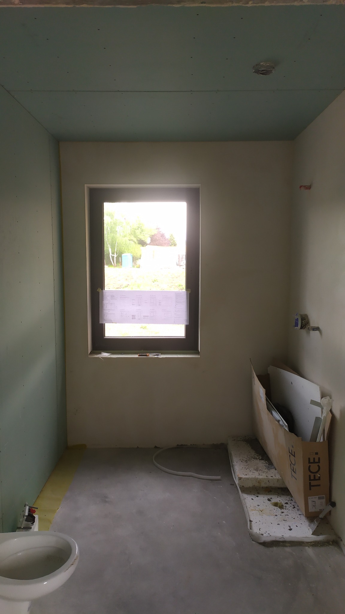 Wnętrze łazienki w trakcie remontu; widoczne płyty gipsowo-kartonowe na ścianach i suficie, surowy beton na podłodze, okno z przyklejonym planem, oraz częściowo zamontowana toaleta.