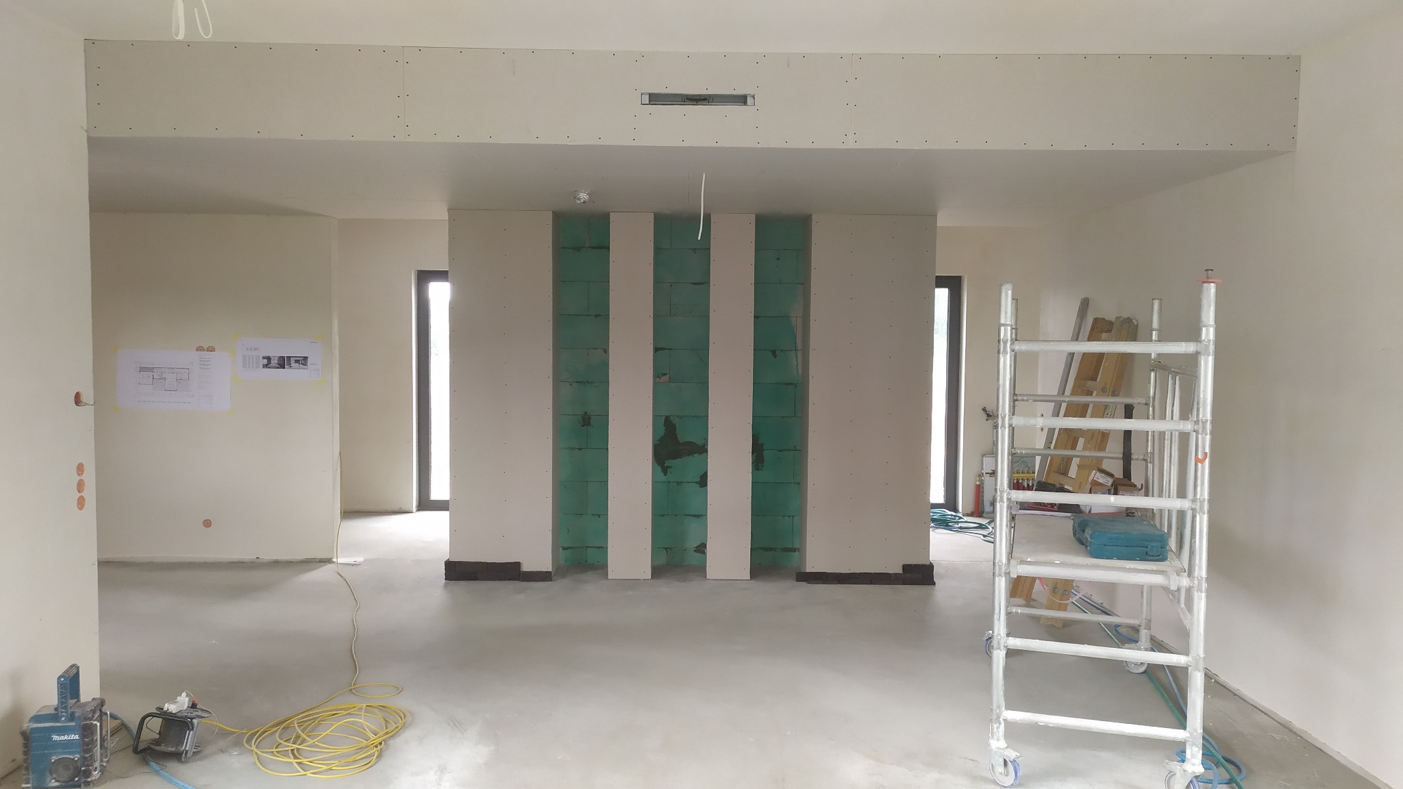 Wnętrze w trakcie prac wykończeniowych: ściana z pionowymi pasami płyt gipsowo-kartonowych na zielonym podłożu, widoczne rusztowanie, szlifierka do gładzi oraz plany na ścianie.