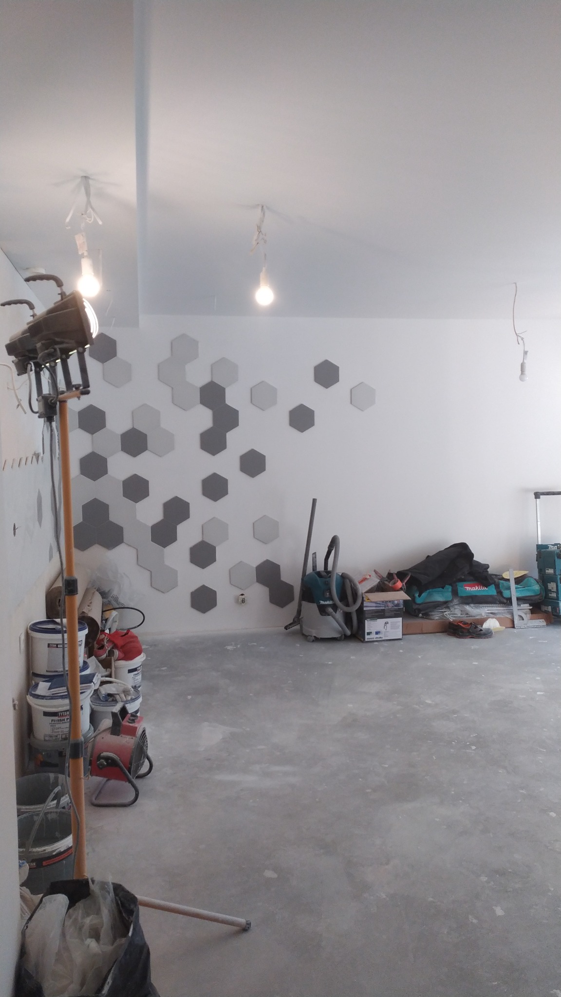 Ściana z szarymi i białymi heksagonalnymi płytkami dekoracyjnymi w trakcie montażu, widoczne narzędzia, puste wiadra po farbie, lampa budowlana i odkurzacz na szarym betonowym podłożu...