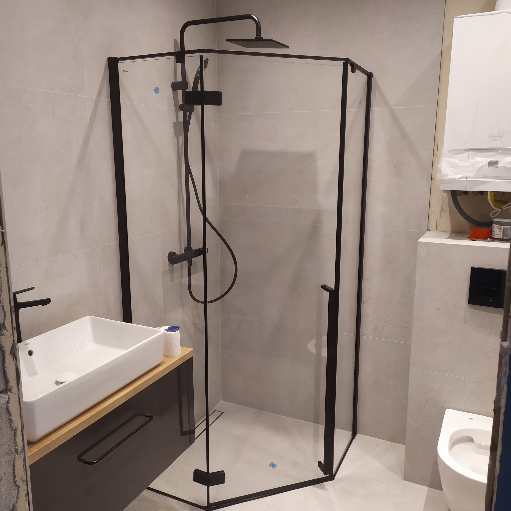 Wykończona łazienka z kabiną prysznicową o czarnych profilach, deszczownicą i baterią w tym samym kolorze, umywalką nablatową na szafce z drewnianym blatem, oraz wiszącą toaletą.