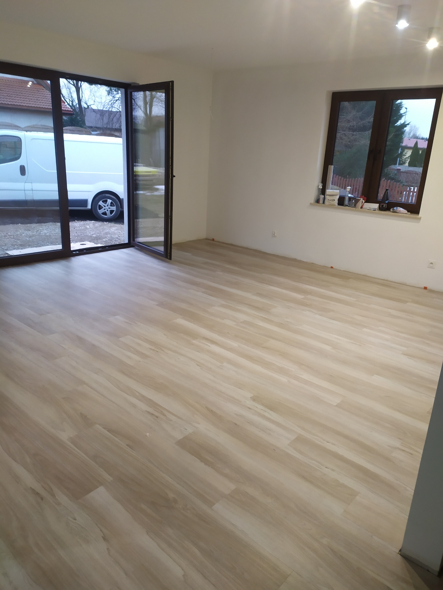 Jasne panele podłogowe ułożone w nowym, pustym pokoju z otwartym balkonowym oknem, przez które widać zaparkowany biały samochód dostawczy.