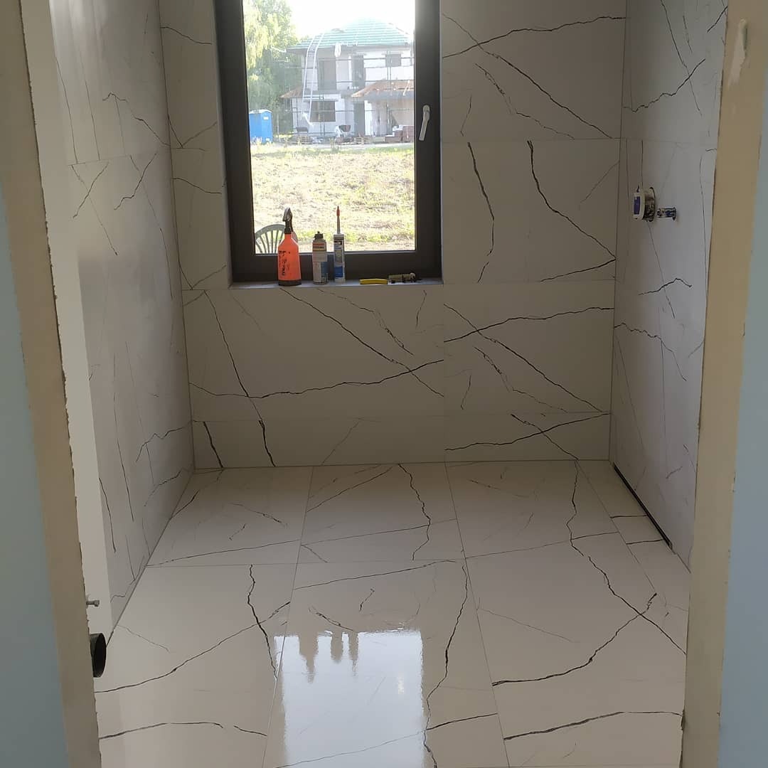 Wykończona łazienka z białymi płytkami imitującymi marmur na ścianach i podłodze, widoczne przybory i narzędzia na parapecie okna, przez które widać budynek w oddali.