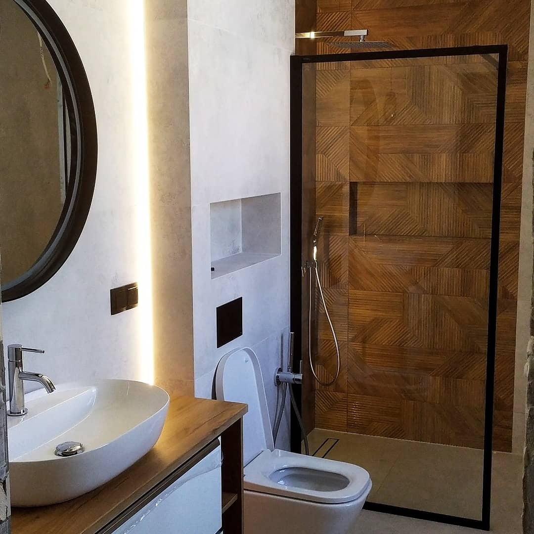 Nowoczesna łazienka z drewnopodobnymi płytkami w kabinie prysznicowej, owalnym lustrem z czarną ramą, umywalką nablatową i podświetleniem LED w ścianie.