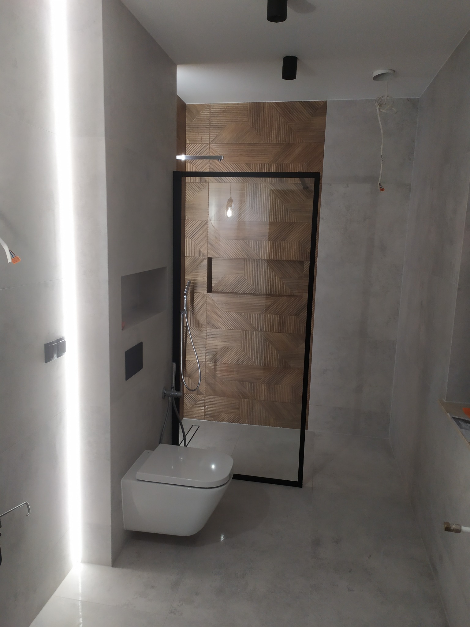 Nowoczesna łazienka z szarymi płytkami, podwieszaną toaletą, kabiną prysznicową z czarną ramą i dekoracyjną ścianą z drewnopodobnych płytek ułożonych we wzór jodełki.