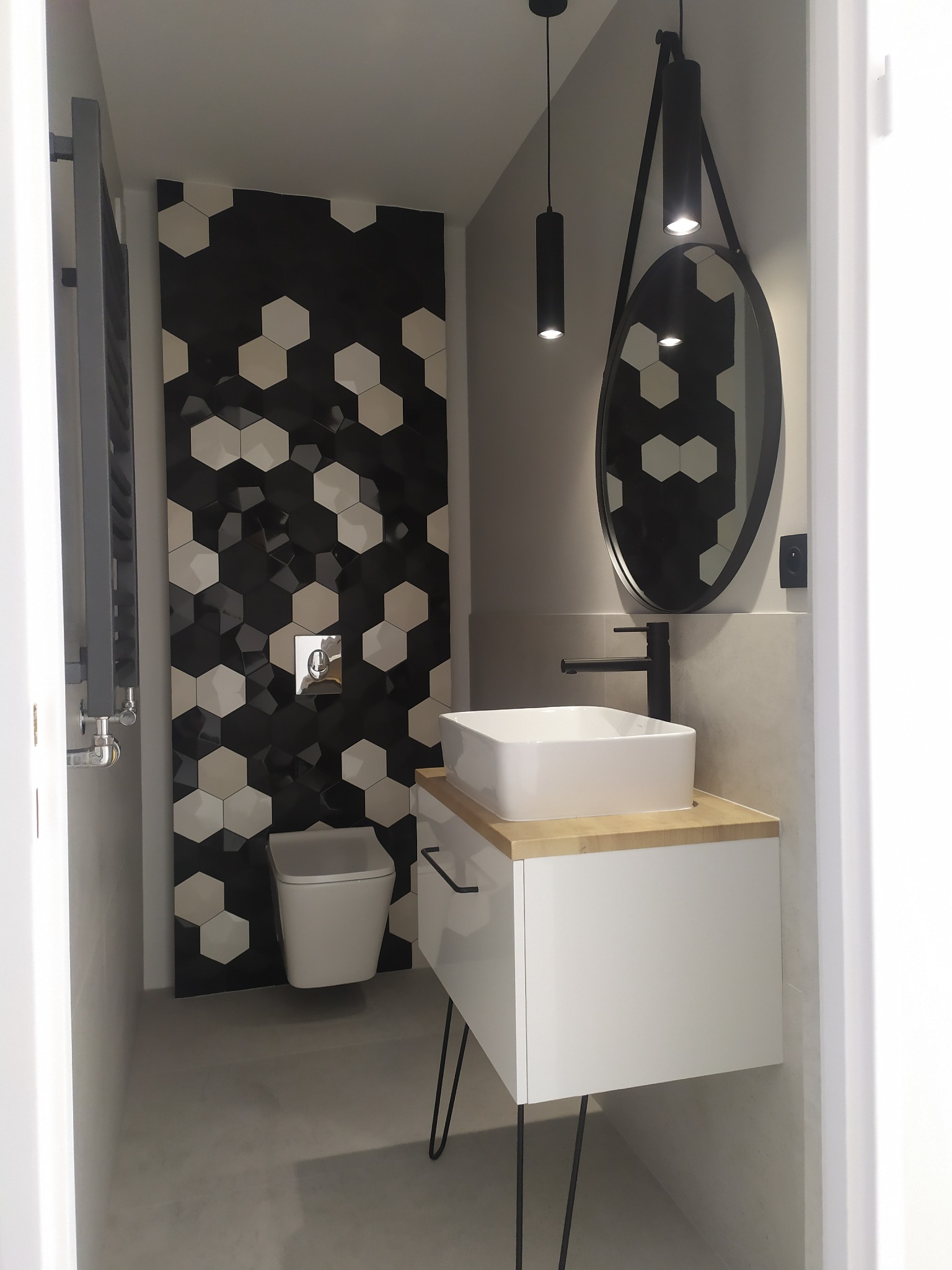 Nowoczesna łazienka z heksagonalnymi czarno-białymi płytkami na ścianie, wisząca umywalka nablatowa z drewnianym blatem, minimalistyczna toaleta i okrągłe lustro na pasku.