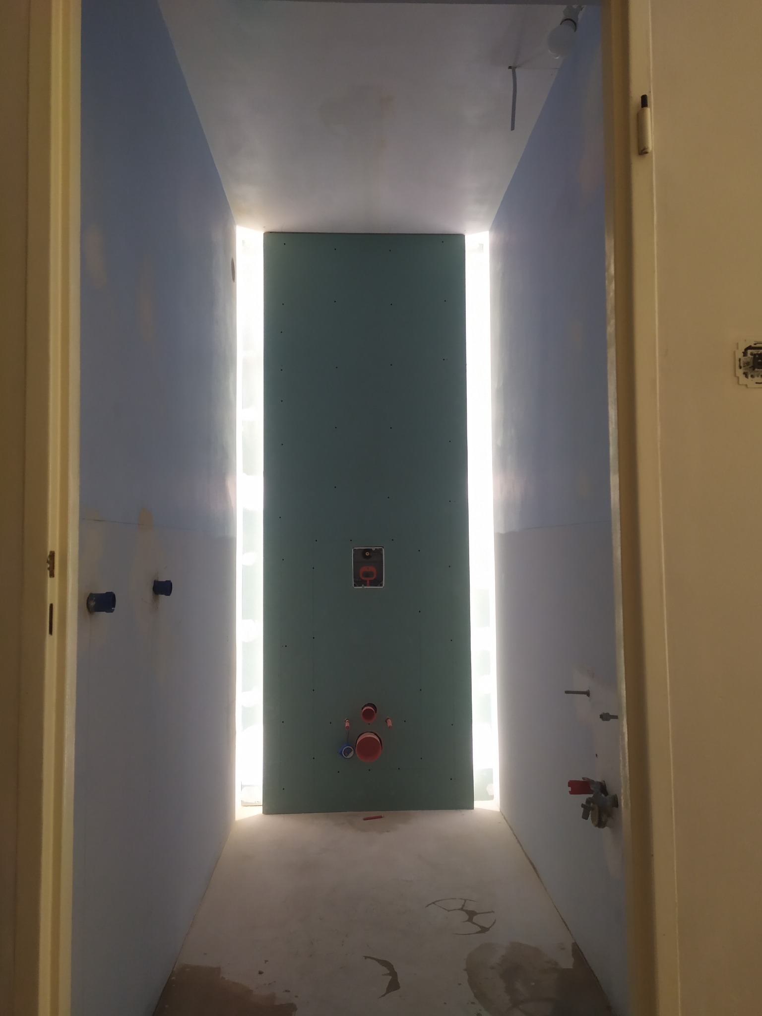 Wnętrze łazienki w trakcie remontu: ściany przygotowane pod gładzie, widoczne instalacje wodne i kanalizacyjne, płyta karton-gips na tylnej ścianie podświetlona od tyłu, surowy beton na podłodze.