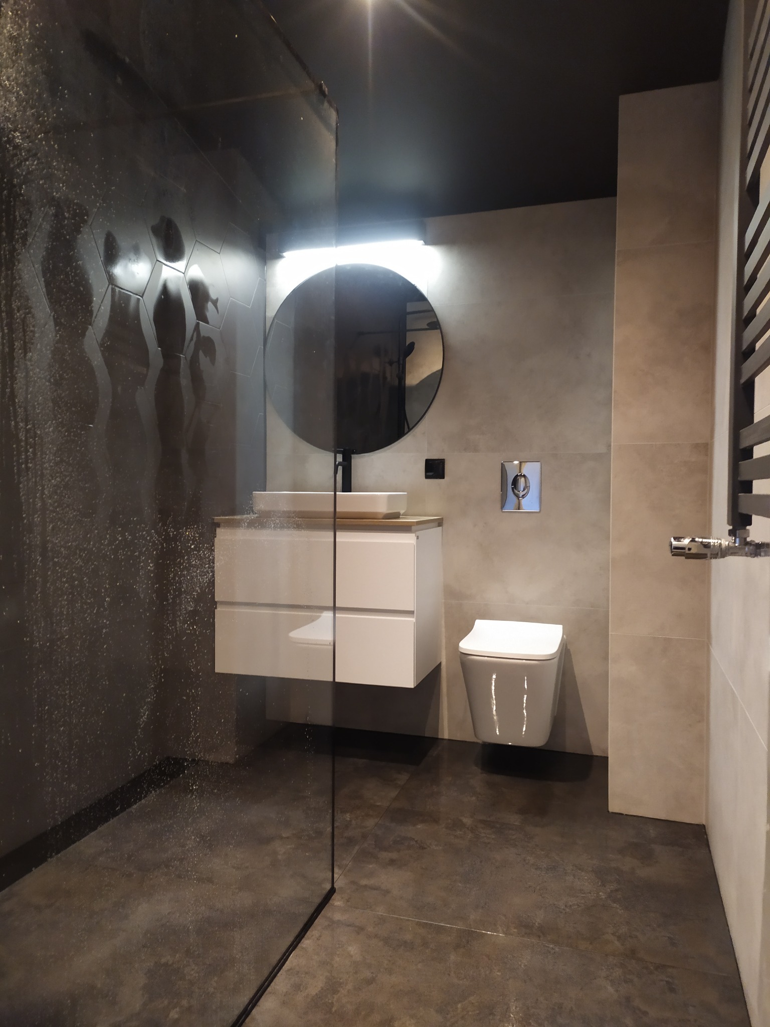 Nowoczesna łazienka z kabiną prysznicową typu walk-in, heksagonalnymi płytkami na ścianie, okrągłym lustrem z podświetleniem, wiszącą szafką z umywalką oraz wiszącą toaletą.