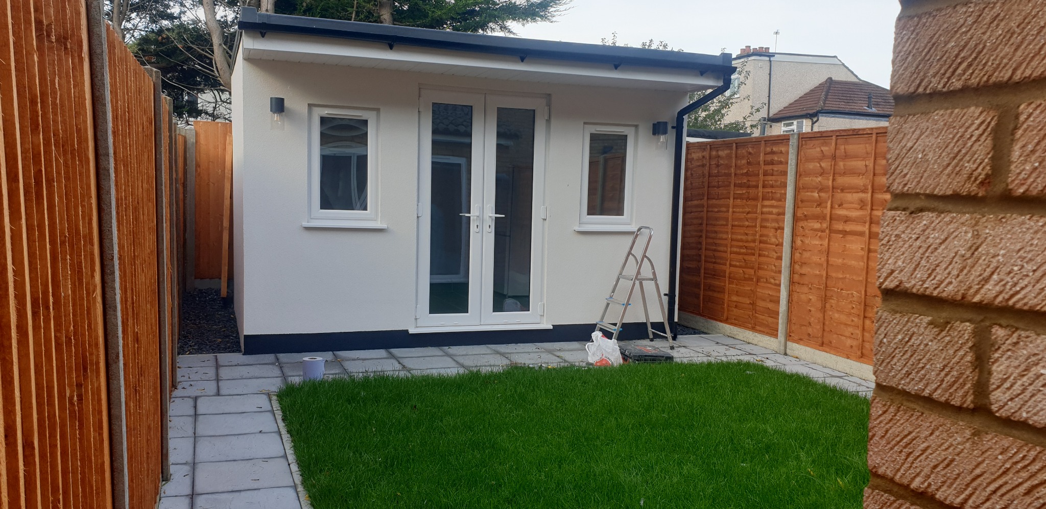 Nowoczesny, biały domek ogrodowy z dwuskrzydłowymi drzwiami i dwoma oknami, wykończony czarnymi detalami, otoczony trawnikiem i drewnianym ogrodzeniem, z drabiną opartą o ścianę.