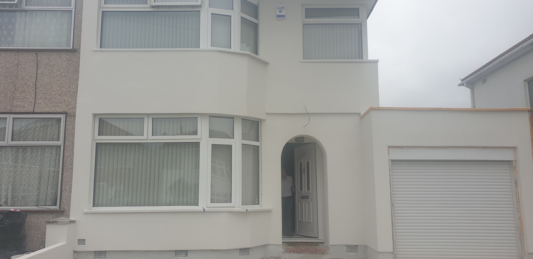 Odnowiona elewacja domu jednorodzinnego z jasną, gładką fakturą, widoczne białe okna z żaluzjami, brama garażowa z roletą i otwarte drzwi wejściowe z numerem 133.