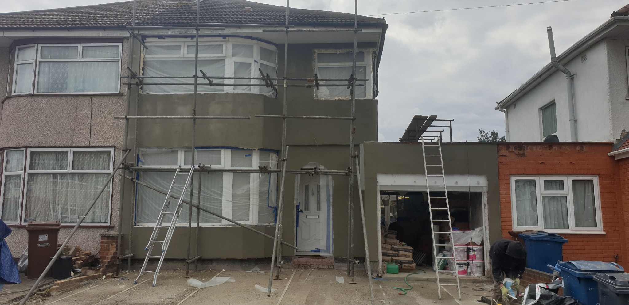 Dom jednorodzinny w trakcie ocieplania elewacji, widoczne rusztowania, okna i drzwi zabezpieczone folią, świeżo nałożona warstwa szpachli, przygotowane worki z materiałem budowlanym w otwartym garażu.