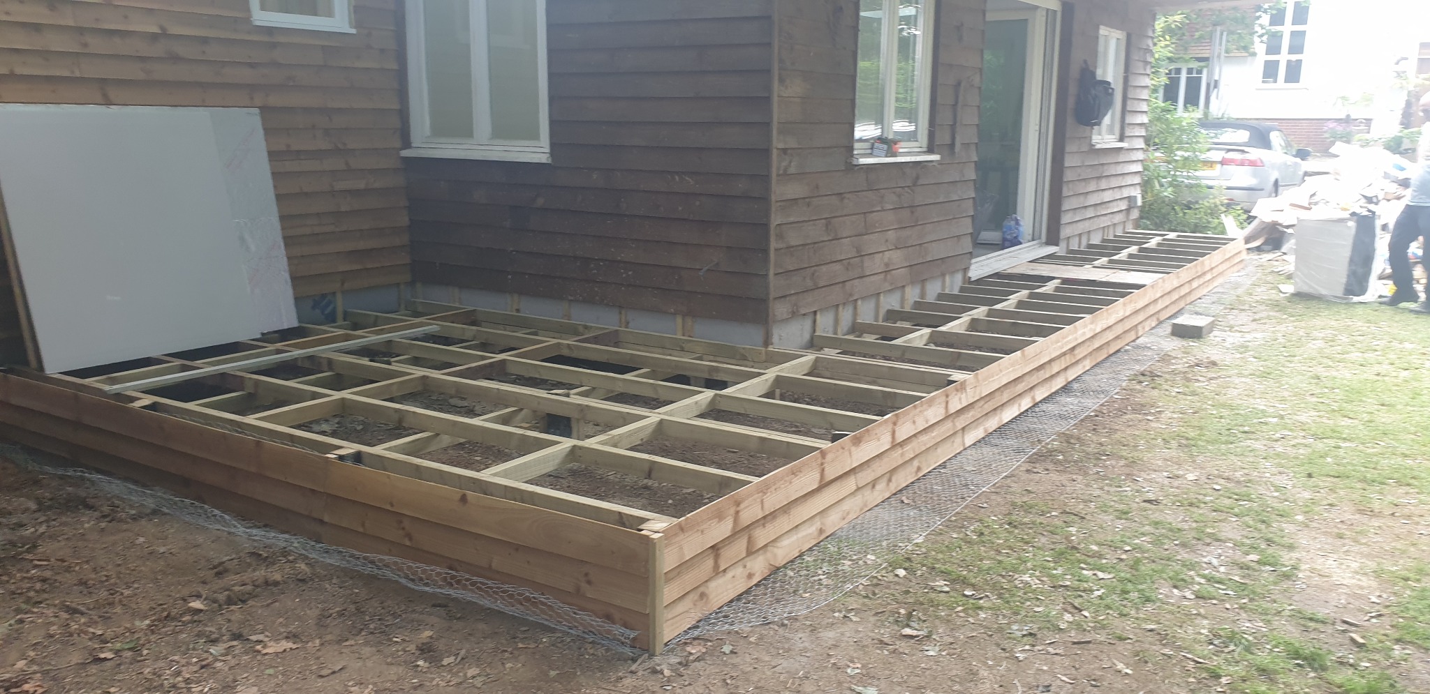 Konstrukcja drewnianego tarasu przylegająca do ściany domu z drewnianą elewacją, zabezpieczona siatką od strony gruntu.