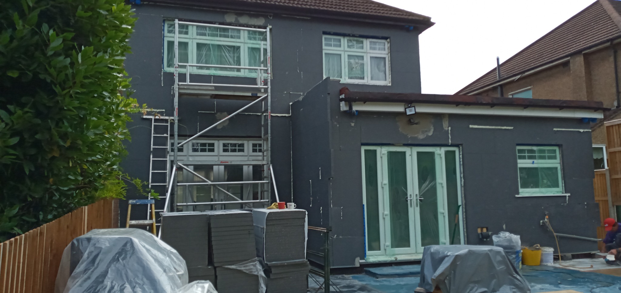 Dom w trakcie przygotowania do ocieplenia elewacji: ściany pokryte warstwą szpachli, okna zabezpieczone folią, rusztowanie ustawione przy oknie, stos płyt izolacyjnych pod folią w pierwszym planie,...