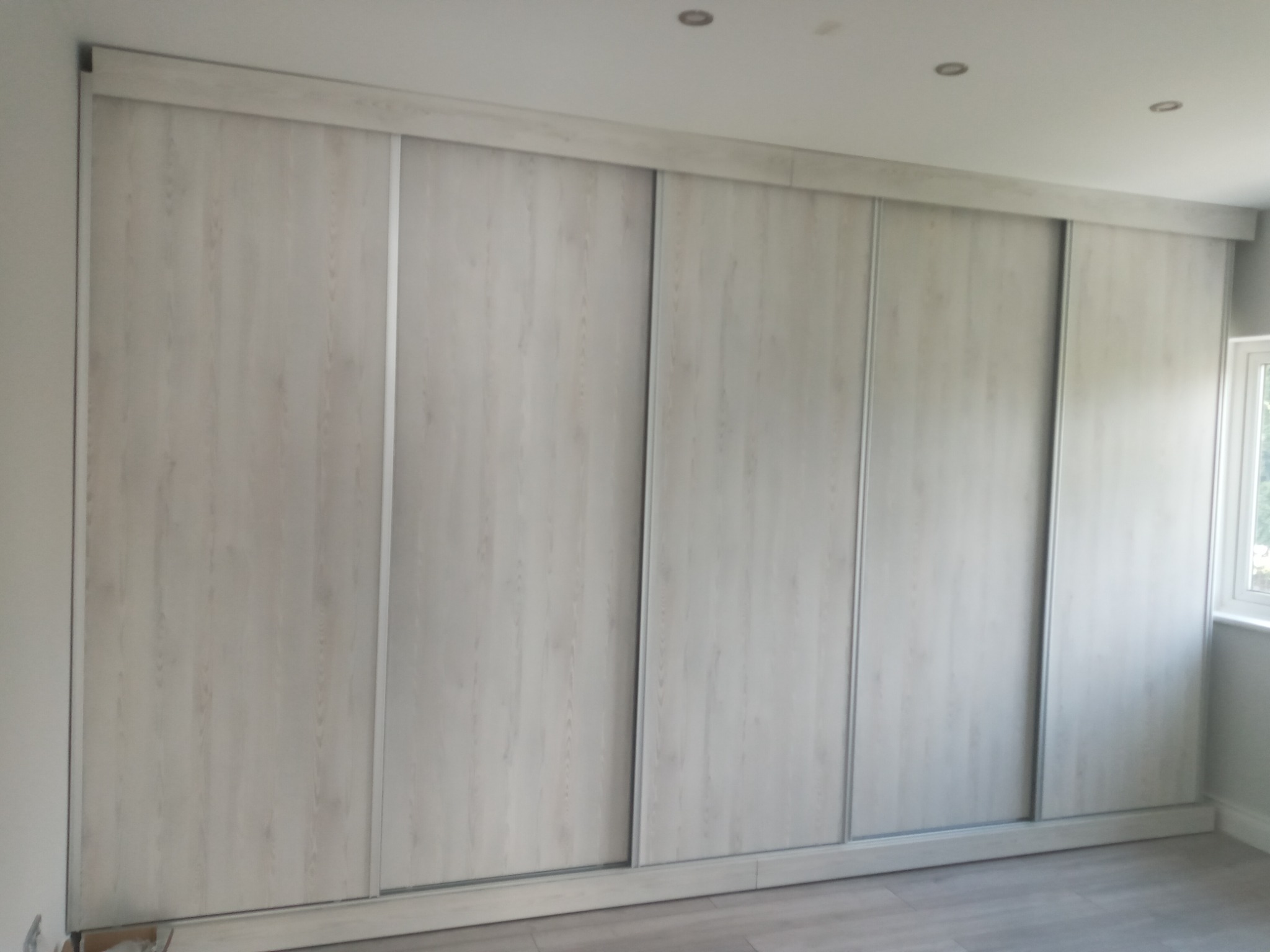 Duża, wbudowana szafa z przesuwnymi drzwiami o jasnym, drewnopodobnym wzorze, zajmująca całą ścianę w pokoju z szarymi panelami podłogowymi i oknem w prawym górnym rogu.