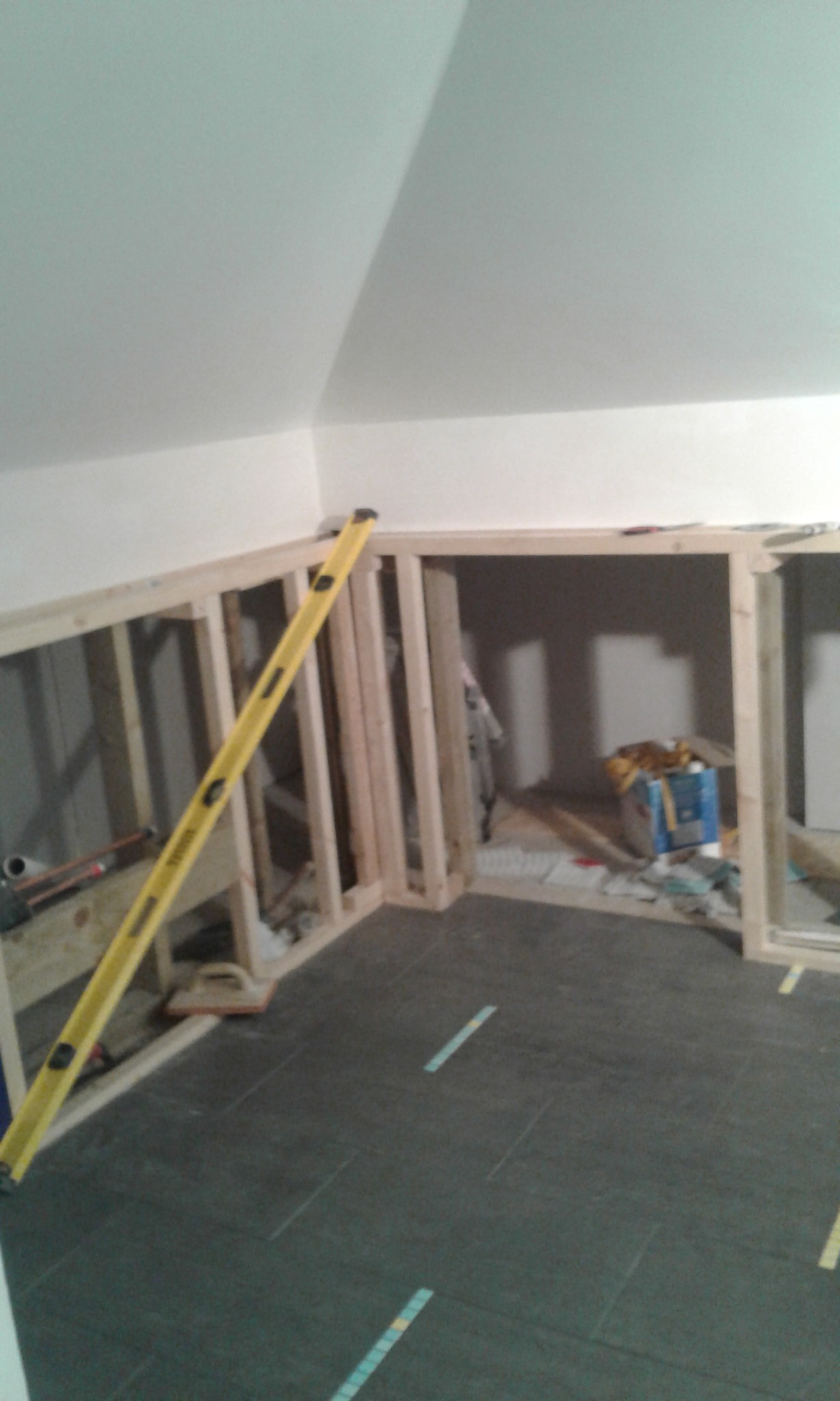 Konstrukcja drewniana ścianki działowej w trakcie budowy, poziomica przyłożona do pionowego słupka, szare płytki podłogowe, widoczne narzędzia i materiały budowlane w tle.