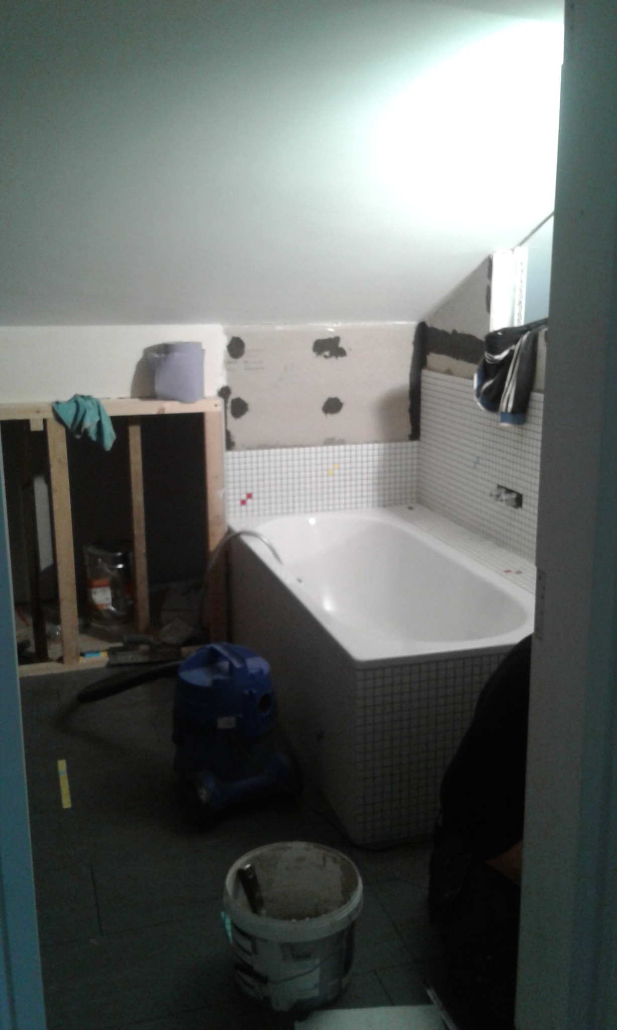 Łazienka w trakcie remontu: biała wanna obłożona mozaiką, ściany przygotowane pod płytki, drewniana konstrukcja w rogu, niebieski odkurzacz przemysłowy i wiadro z zaprawą na podłodze.