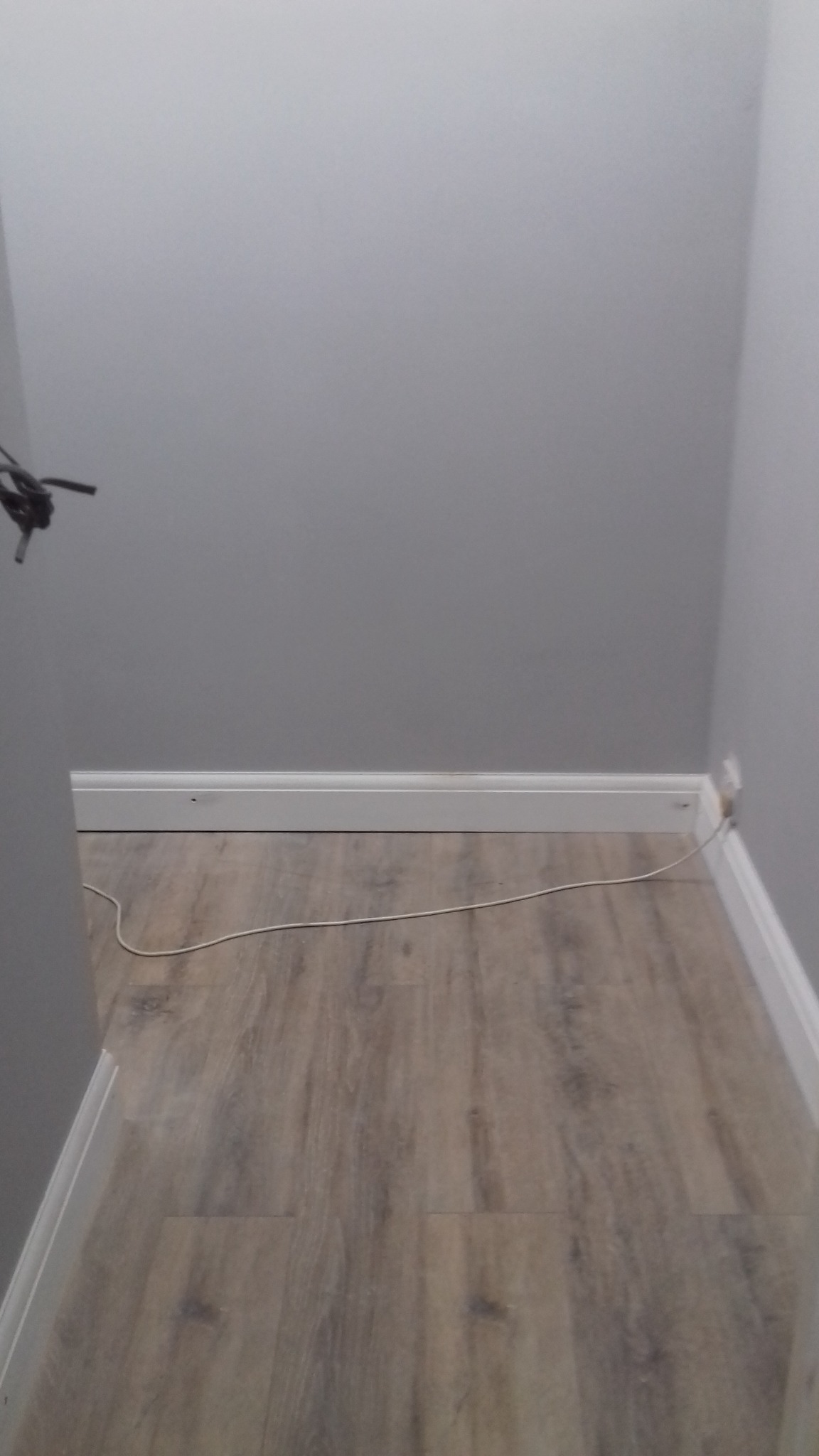 Wąski korytarz z szarą ścianą, panelami podłogowymi imitującymi drewno i białą listwą przypodłogową. Widoczny kabel elektryczny biegnący wzdłuż ściany.