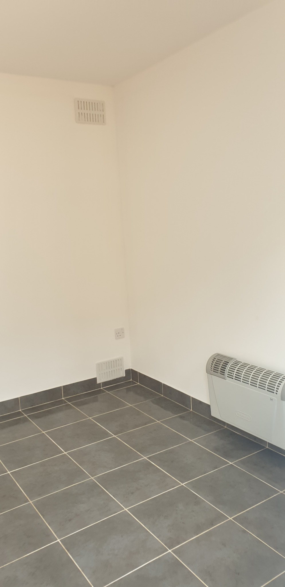 Pusty pokój po remoncie z szarymi płytkami podłogowymi, grzejnikiem elektrycznym i kratkami wentylacyjnymi w ścianie.
