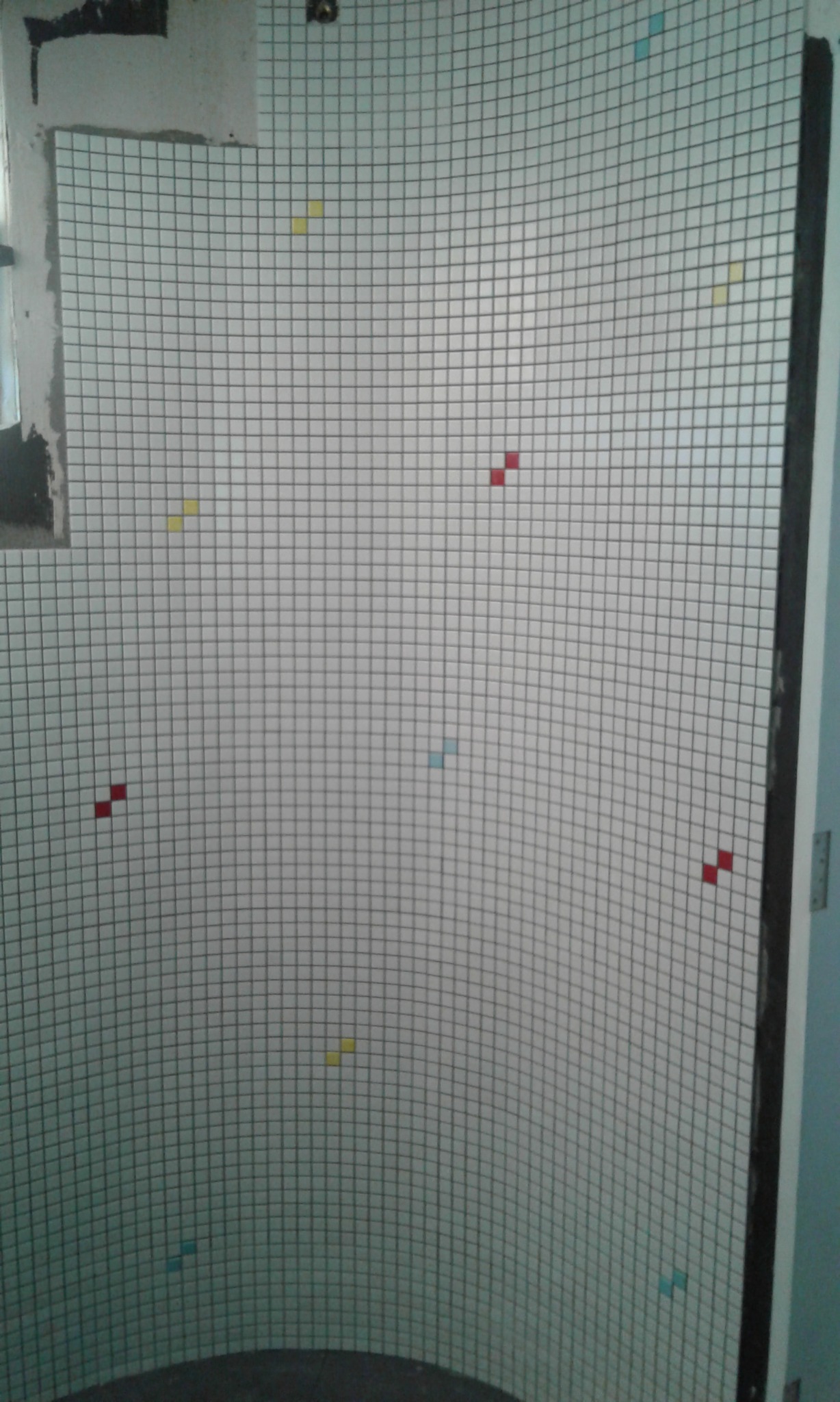 Ściana łazienki w trakcie układania mozaiki z drobnymi, kolorowymi akcentami na siatce, widoczny fragment okna i niedokończone wykończenie ściany.