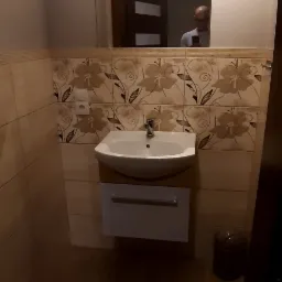 Umywalka z szafką podwieszaną w łazience, ściany wyłożone płytkami z motywem kwiatowym, lustro z odbiciem osoby, drewnopodobna podłoga.