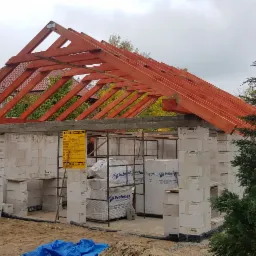 Wznoszenie ścian budynku z bloczków betonowych z zamontowaną drewnianą konstrukcją dachu, widoczna tablica informacyjna i materiały budowlane na placu budowy.