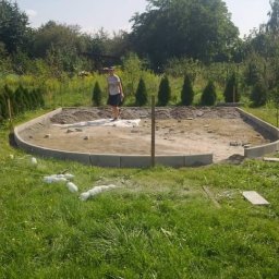 Arkas - Przygotowanie podłoża pod taras lub ścieżkę z obrzeżami z betonowej kostki, widoczna osoba na placu budowy i butelki po napojach na trawie.