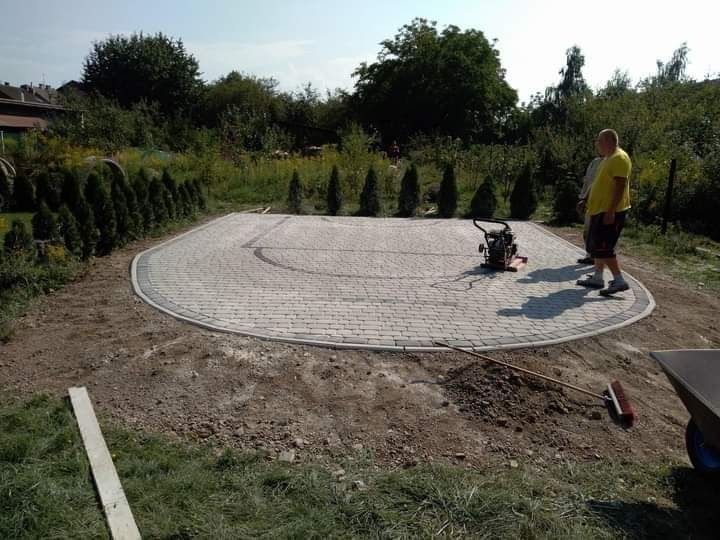 Półokrągły podjazd z jasnoszarej kostki brukowej, obrzeża z ciemniejszej kostki, zagęszczarka w tle, widoczne narzędzia i dwóch pracowników, trawnik i tuje w tle.
