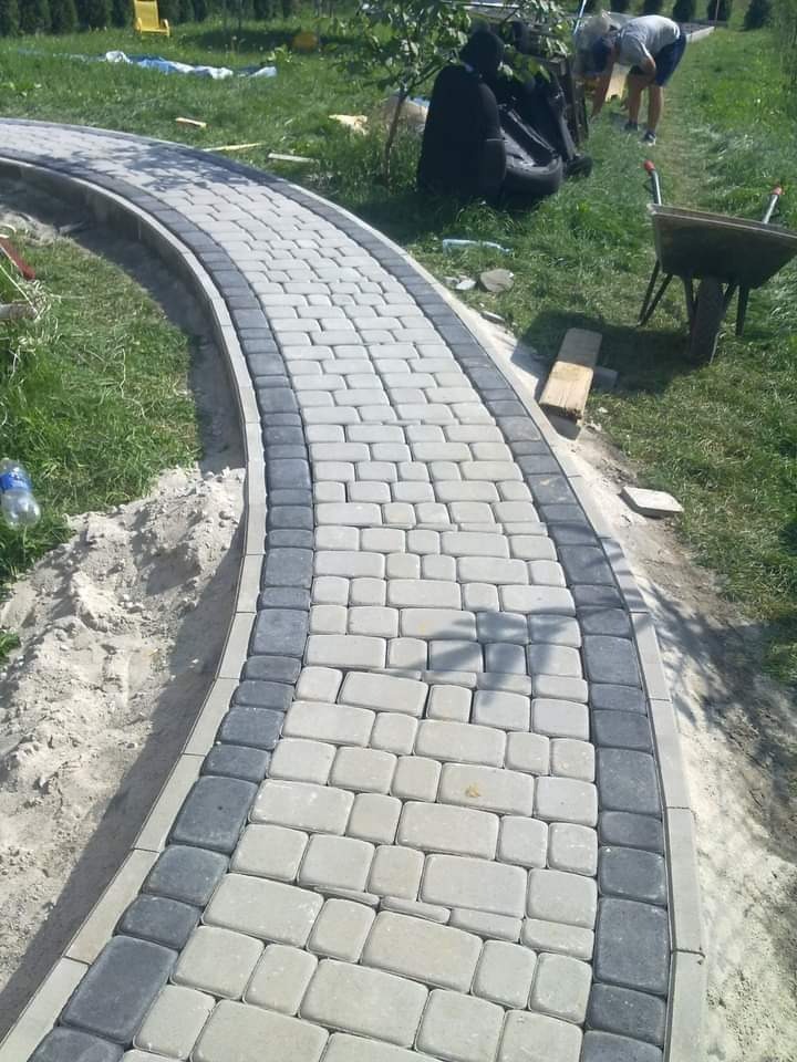 Zakrzywiony chodnik z jasnoszarej kostki brukowej z ciemnoszarym obramowaniem, w tle trawnik i osoby pracujące przy wózku.