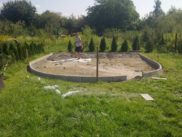 Przygotowanie podłoża pod taras lub ścieżkę z obrzeżami z betonowej kostki, widoczna osoba na placu budowy i butelki po napojach na trawie.