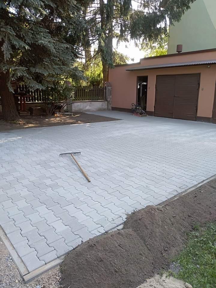 Ułożona szara kostka brukowa typu 'psi pazur' na podjeździe, widoczny grabie i ubijarka w tle, świeżo obsypana ziemią krawędź.