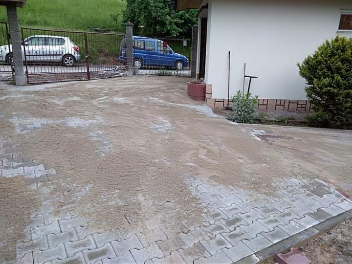 Przygotowanie podłoża z piasku pod ułożenie szarej kostki brukowej typu 'kość' na podjeździe, widoczne częściowo ułożone fragmenty, w tle brama wjazdowa i zaparkowane samochody.