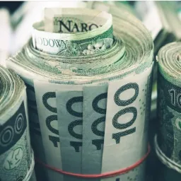 Zwinięte pliki banknot&oacute;w 100 PLN spięte czerwoną gumką, widoczne detale nominał&oacute;w i zabezpieczeń.