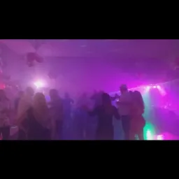 Tłum ludzi tańczących w zadymionym pomieszczeniu oświetlonym fioletowymi i zielonymi światłami podczas imprezy.
