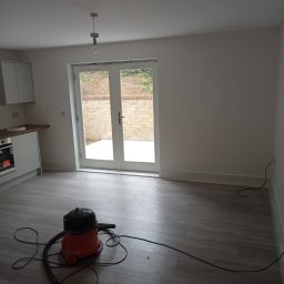 Prywatny wykonawca - Wnętrze po remoncie z ułożonymi panelami podłogowymi w odcieniach szarości, widoczny odkurzacz przemysłowy i białe ściany, drzwi balkonowe z widokiem na ogród.