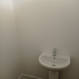 Prywatny wykonawca - Biała umywalka na postumencie z chromowaną baterią w narożnej łazience z pomalowanymi na biało ścianami i ciemną podłogą.