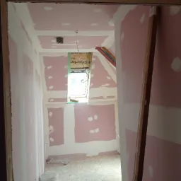 Poddasze w trakcie adaptacji: płyty gipsowo-kartonowe na ścianach i suficie, częściowo pokryte białą warstwą, okno dachowe z widoczną konstrukcją, dwa białe wiadra z farbą na podłodze z płyty OSB.