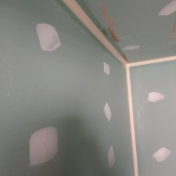 Prywatny wykonawca - Ściana pomalowana na zielono z widocznymi plamami po szpachlowaniu, przygotowana do dalszych prac wykończeniowych. Widoczne narożniki zabezpieczone taśmą malarską.