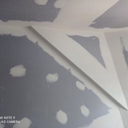 Prywatny wykonawca - Ściana i sufit w trakcie prac wykończeniowych, widoczne fragmenty szpachli i biała listwa dekoracyjna pod sufitem, na ścianie widoczne punktowe naprawy szpachlą.