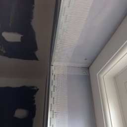 Prywatny wykonawca - Przygotowanie do wyrównywania ściany przy ościeżnicy: siatka wzmacniająca nałożona na narożnik, widoczne ślady po wcześniejszych pracach na ścianie obok.