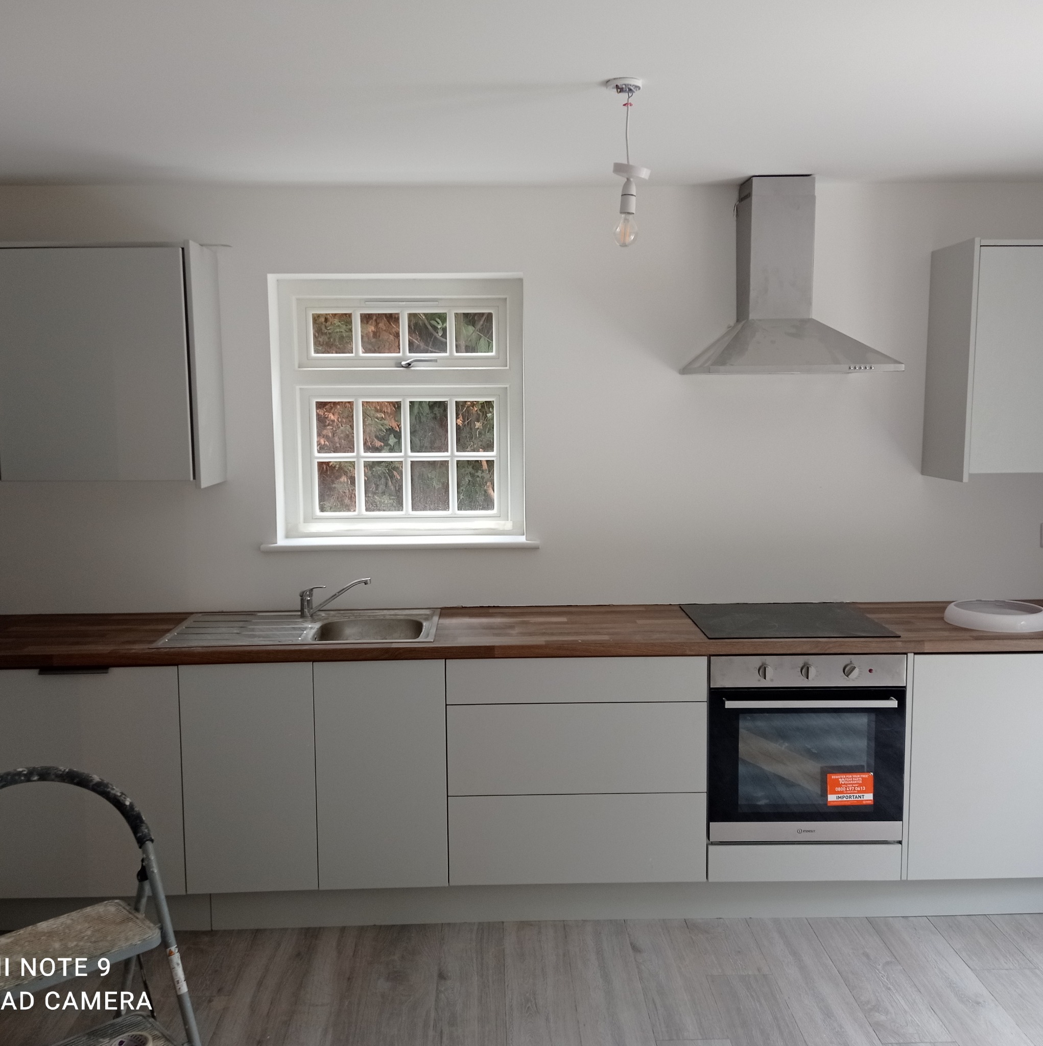 Jasna kuchnia z szarymi szafkami i drewnianym blatem, widok na okno z zielenią na zewnątrz, drabina malarska w lewym dolnym rogu, surowa żarówka zwisająca z sufitu, srebrny okap nad płytą kuchenną.