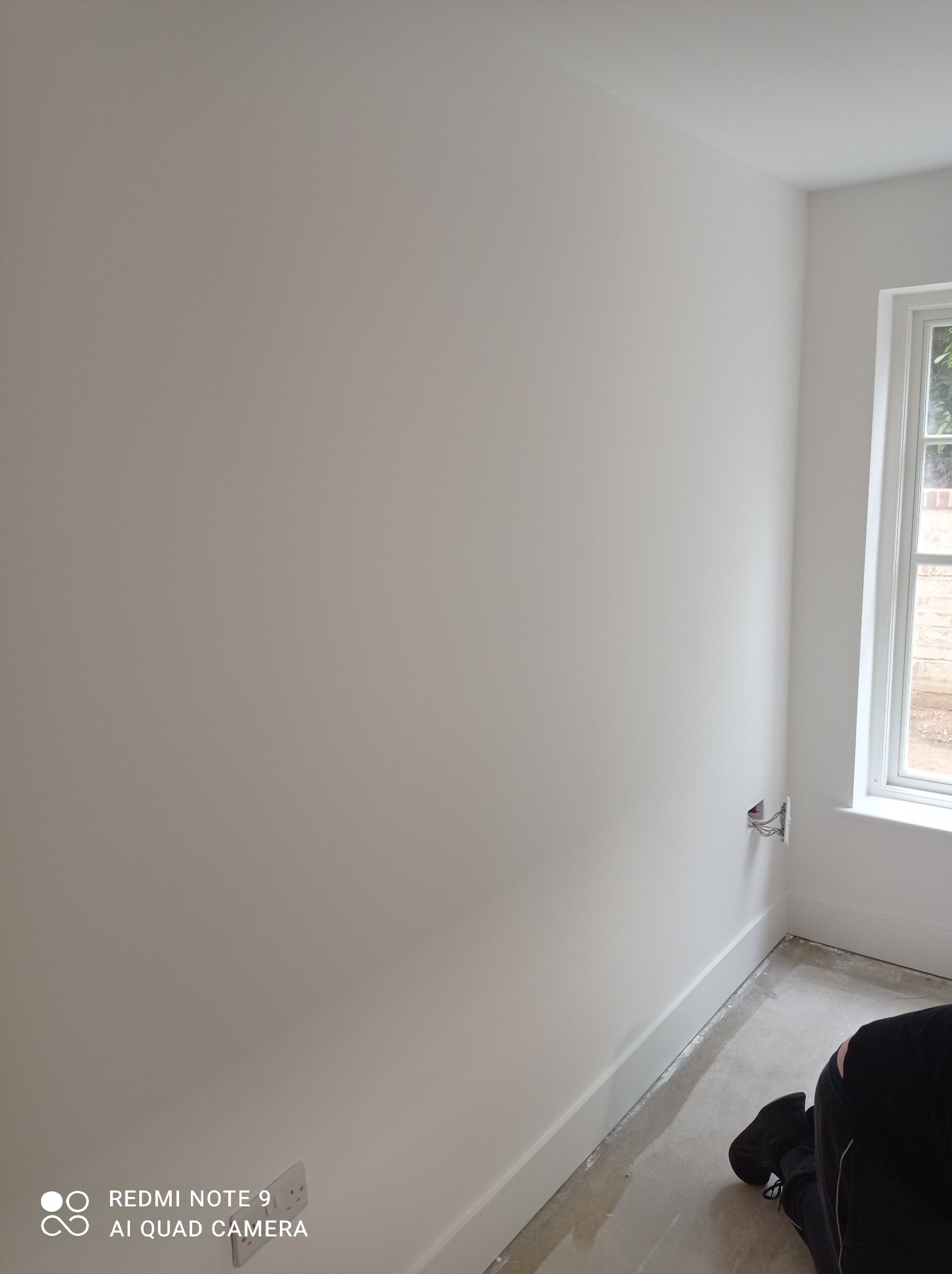 Świeżo pomalowana na biało ściana w rogu pokoju z widocznym gniazdkiem elektrycznym z wystającymi kablami, białą listwą przypodłogową i fragmentem okna z widokiem na zewnątrz, podłoga przygotowana...