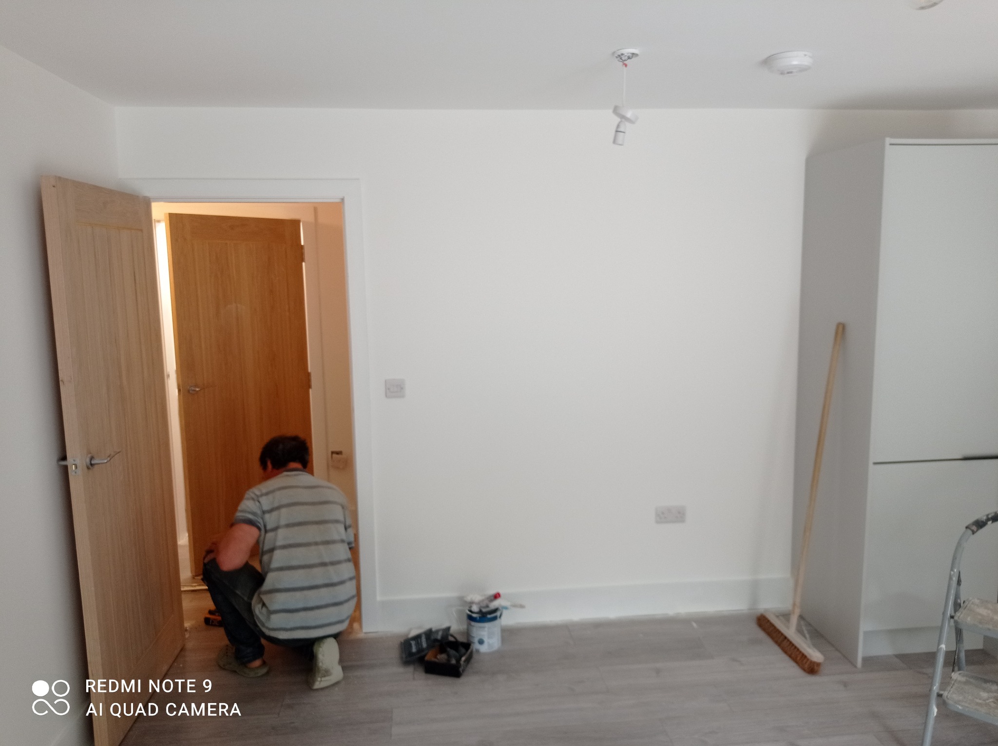 Mężczyzna w paski, w kucki, przy futrynie drzwi, w trakcie prac wykończeniowych w jasnym pomieszczeniu z panelami podłogowymi; widoczne narzędzia malarskie, puszka z farbą, listwa przypodłogowa...