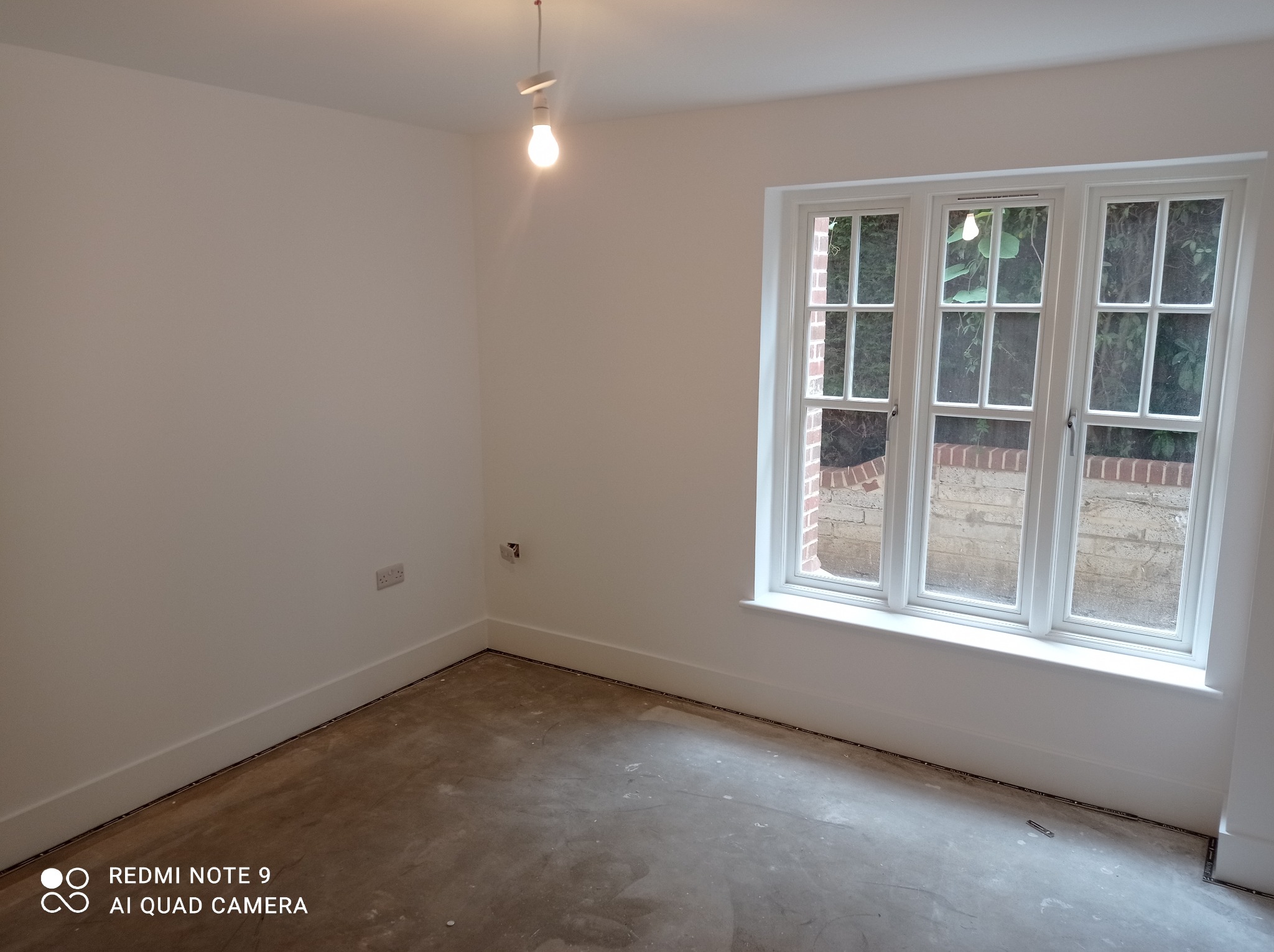 Wnętrze pokoju po malowaniu ścian na biało, widoczne gładkie ściany, okno z białymi ramami i surowa podłoga przygotowana do dalszych prac wykończeniowych.