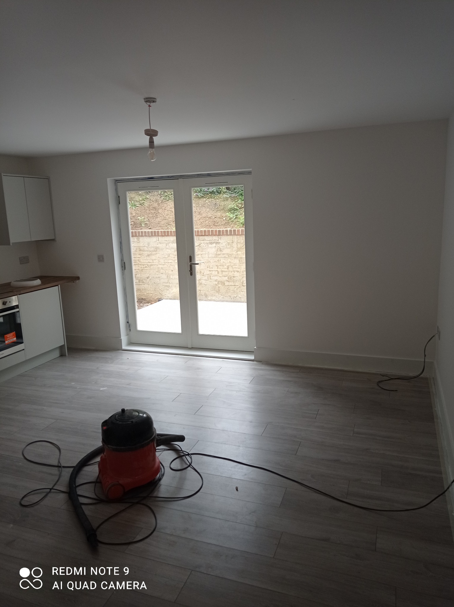 Wnętrze po remoncie z ułożonymi panelami podłogowymi w odcieniach szarości, widoczny odkurzacz przemysłowy i białe ściany, drzwi balkonowe z widokiem na ogród.