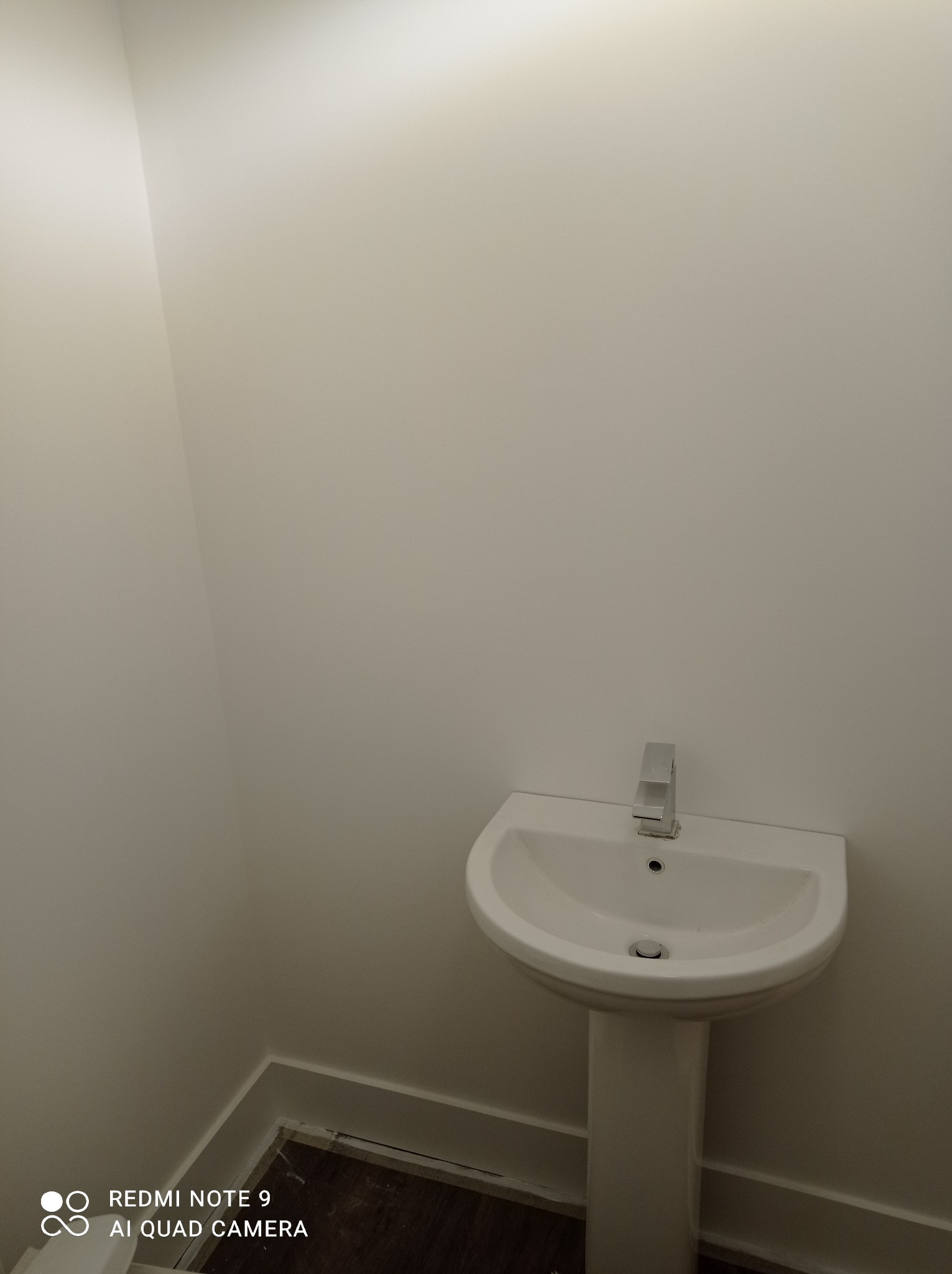 Biała umywalka na postumencie z chromowaną baterią w narożnej łazience z pomalowanymi na biało ścianami i ciemną podłogą.